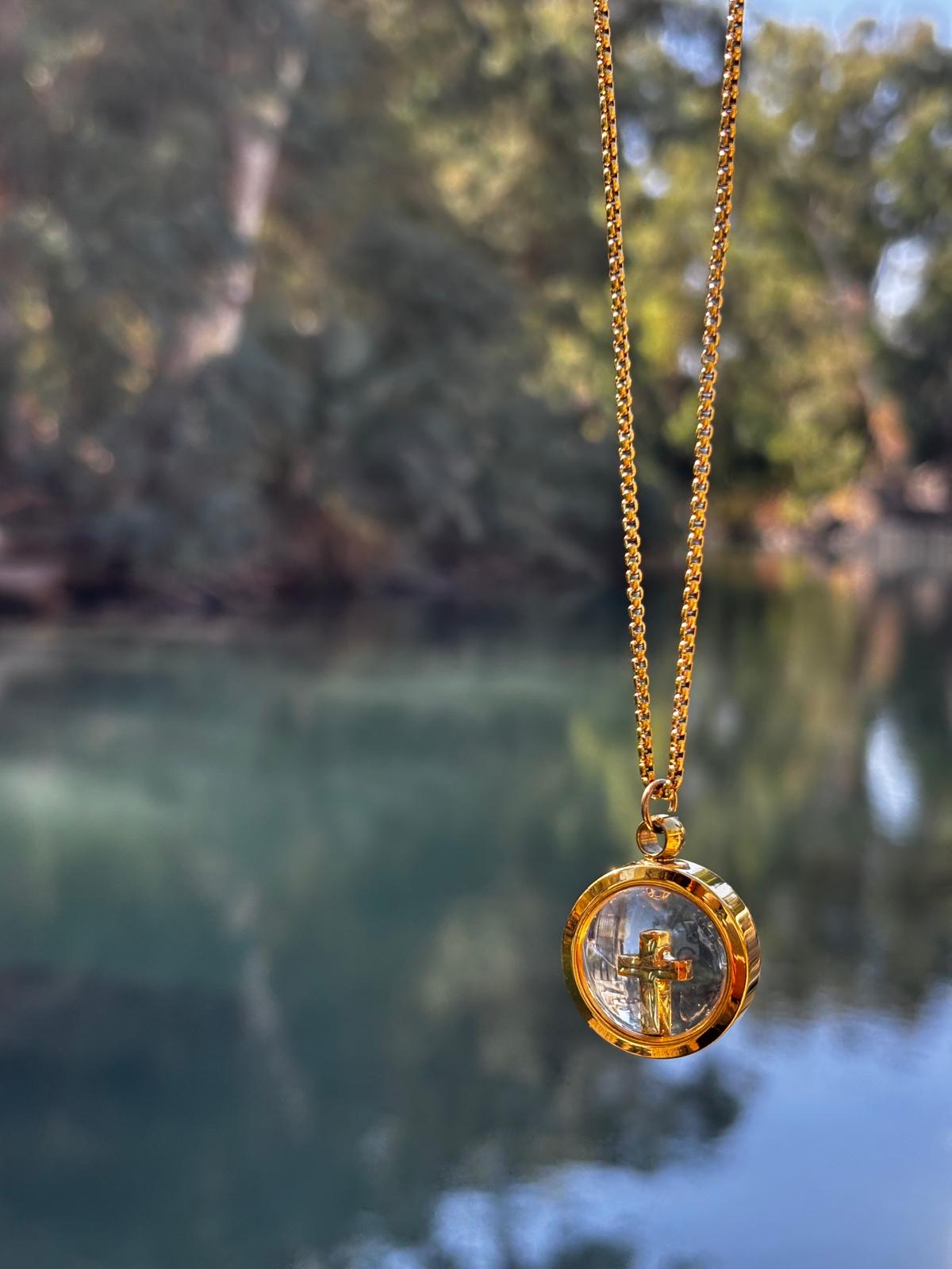 Gold necklace with a clear pendant featuring a cross, against a blurred natural background.