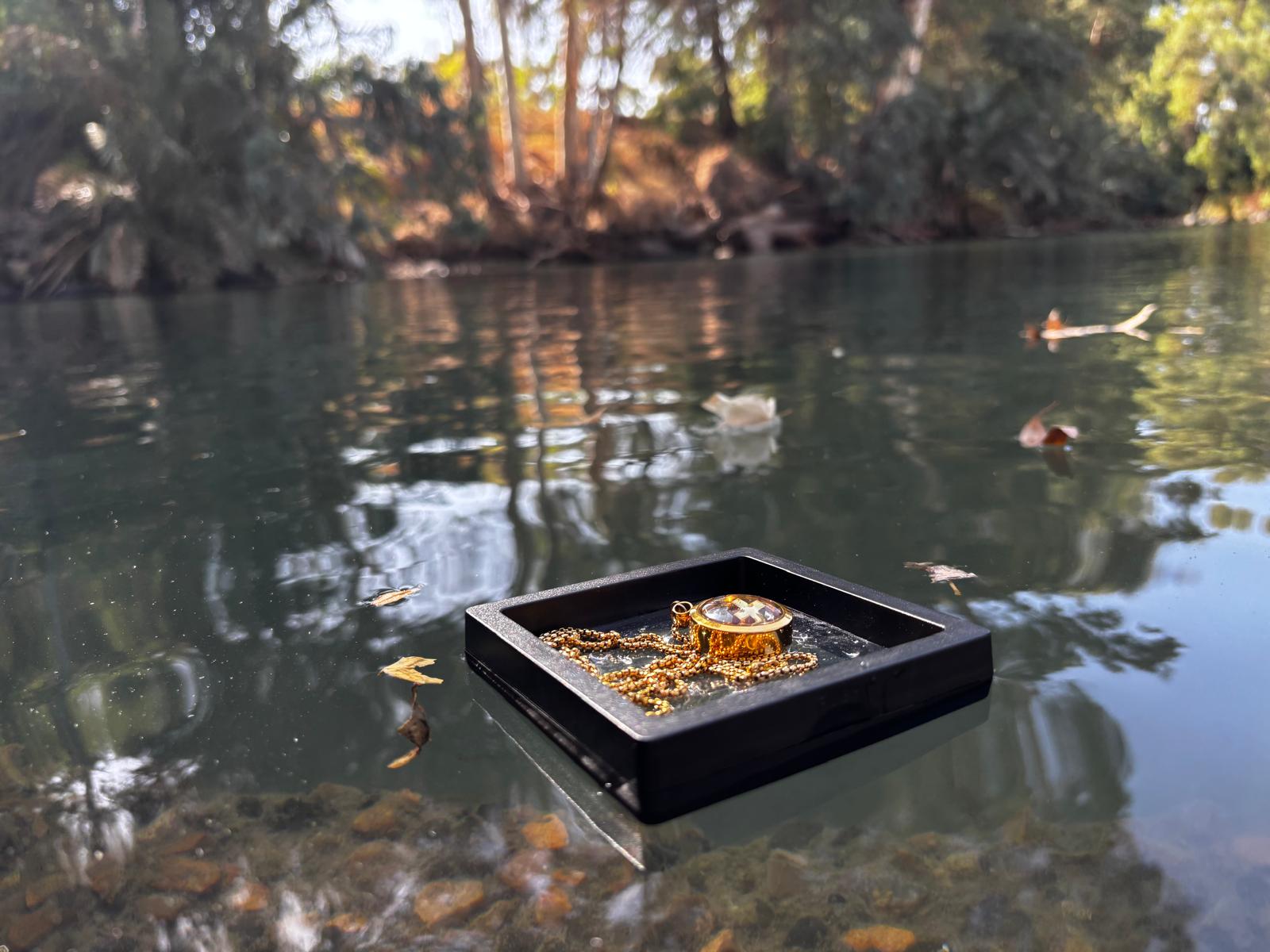 Black jewelry display case with gold necklace on a reflective water surface with trees in the background