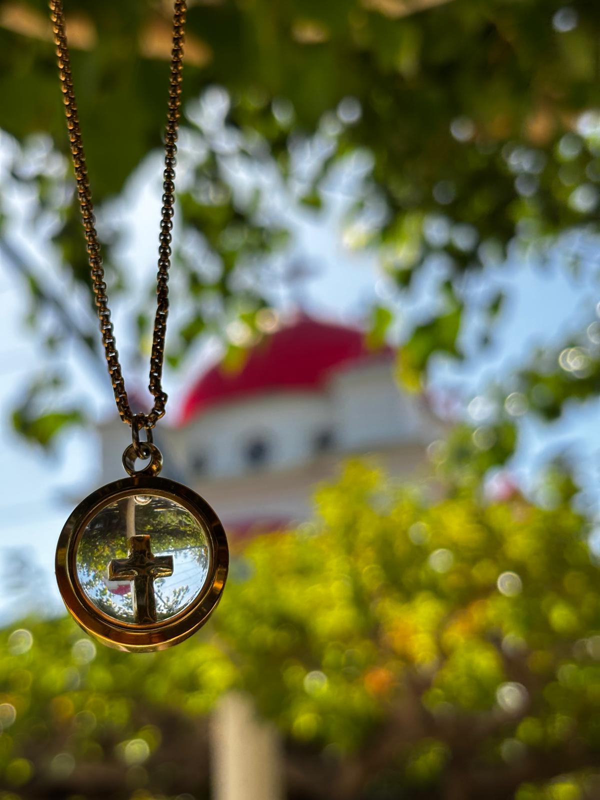 Necklace with a circular pendant featuring a cross, blurred green foliage and building in the background