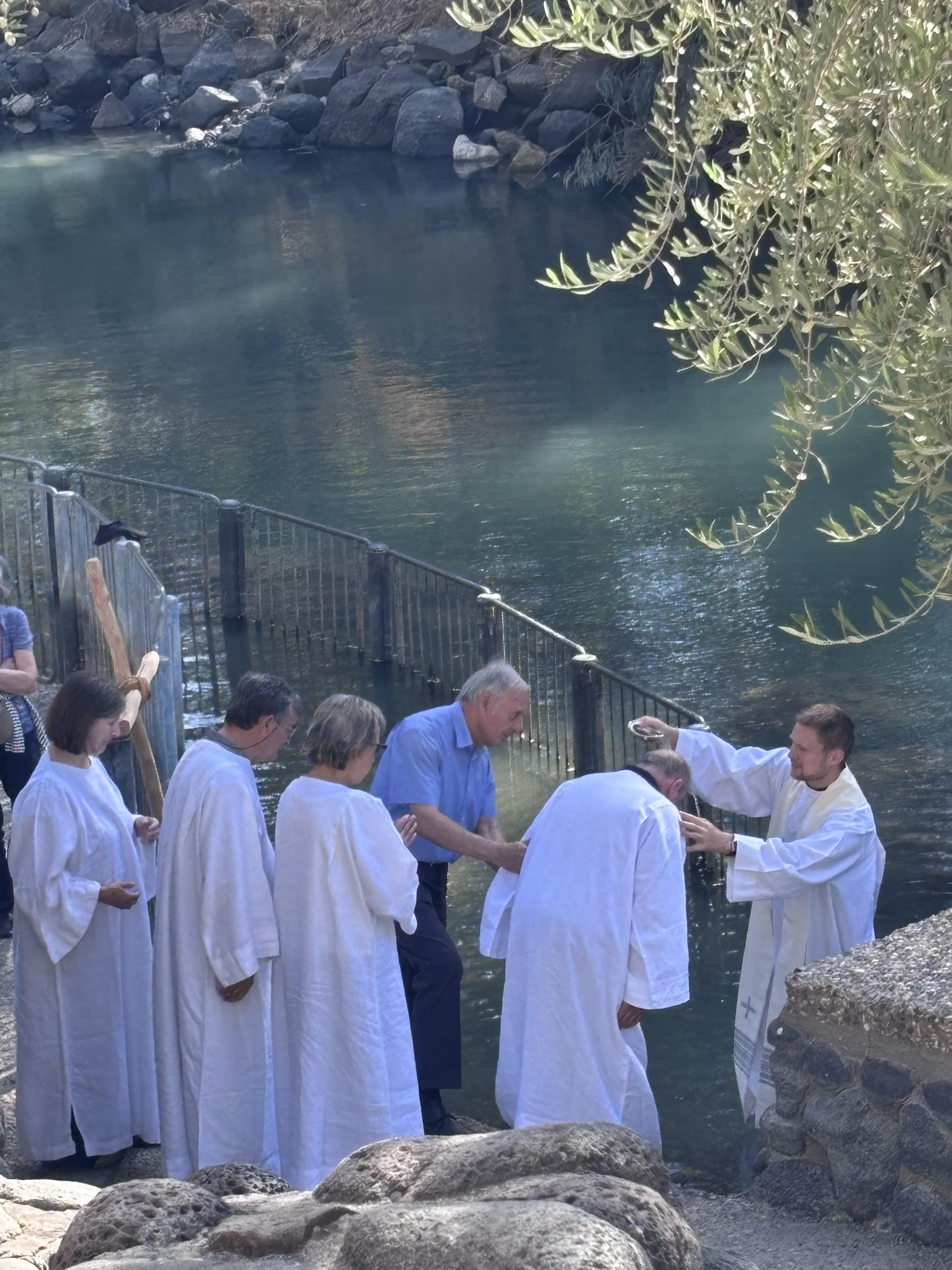 Group of people in white robes by a body of water, possibly participating in a religious ceremony.